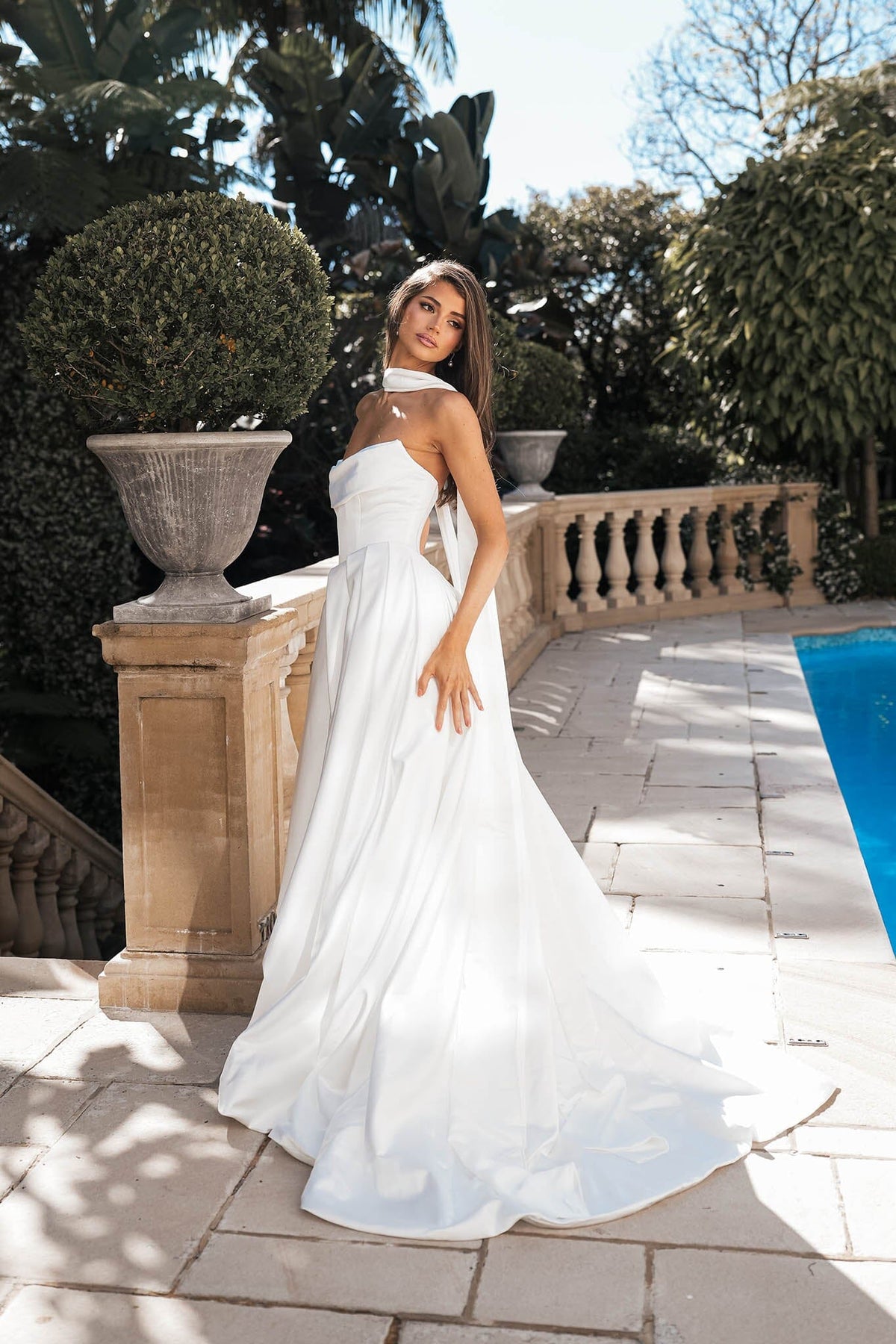 Modern bride wearing white strapless ballgown wedding dress with a Basque waist, draped neckline and leg slit styled with long white satin scarf