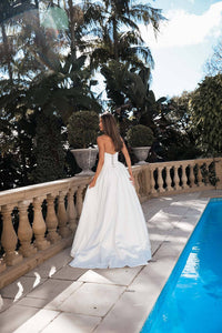 Modern bride wearing white strapless ballgown wedding dress with a Basque waist, draped neckline and leg slit walking along poolside