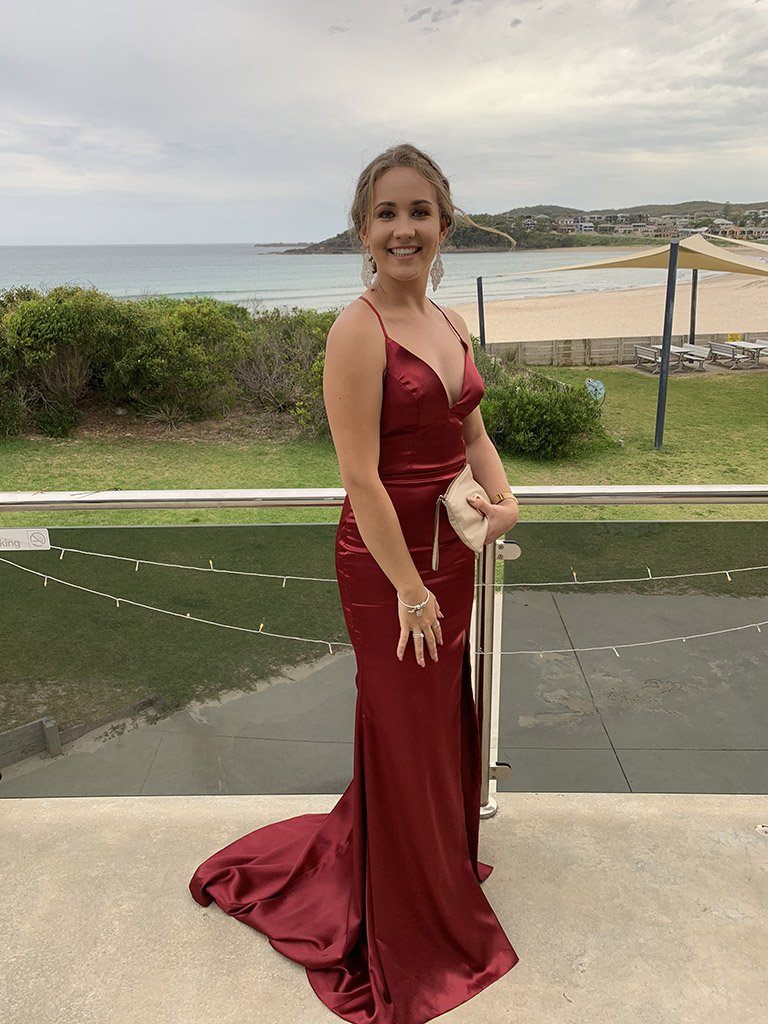 Noodz Boutique's customer wearing Electra sleeveless satin evening gown with deep v neckline, lace up back design, front left high split and long train